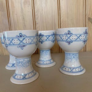 May include: A set of three white ceramic egg cups featuring a blue painted pattern. Each cup has a wide base, a rounded bowl, and a decorative band of blue cross-hatching and dots around the centre, with a speckled blue design on the stem.