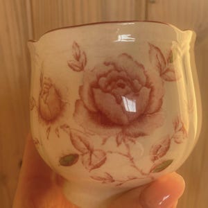 May include: A cream-coloured ceramic sugar bowl or small vessel featuring a delicate rose pattern in a muted reddish-brown tone. The surface displays a subtle crackle glaze finish, and the rim has a gentle, scalloped edge design.