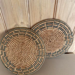 May include: Two circular woven wall baskets featuring a concentric spiral pattern. The baskets are constructed from natural tan and muted sage green fibres, creating a repeating alternating colour design throughout the flat, round woven surfaces.