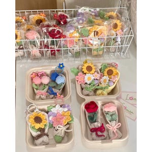 May include: A collection of handmade crochet flower bouquets in various colors, including sunflowers, roses, and daisies. Some bouquets are packaged in clear plastic, while others are displayed in beige containers. Keychains are attached to some bouquets.