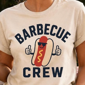Può includere: Una maglietta beige chiaro con la scritta "BARBECUE CREW" e un cartone animato di un hot dog con occhiali da sole. L'immagine mostra una donna che tiene in mano una bottiglia di birra vicino a una griglia con hamburger. Un cartello recita "GOOD FOOD, GOOD FRIENDS, GOOD TIMES."