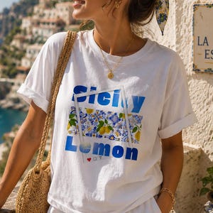 May include: A woman wearing a white t-shirt and shorts with a straw bag. The t-shirt has the words "Sicily Lemon" in blue, with a lemon and floral design. The background shows a coastal town and the sea.