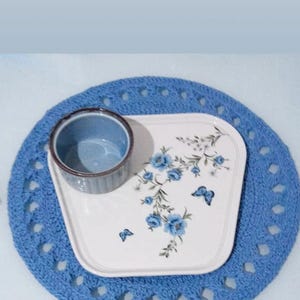 May include: A blue crocheted placemat with a white rectangular plate featuring blue floral and butterfly designs. A small blue bowl sits on the plate. The text "@sizdengelenler" is visible.