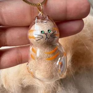 Puede incluir: Un colgante de gato de resina transparente con rayas naranjas y ojos verdes, suspendido de un cordón dorado. El colgante está relleno de pelo marrón claro, lo que le da una textura única. La cara del gato está pintada a mano con bigotes negros.