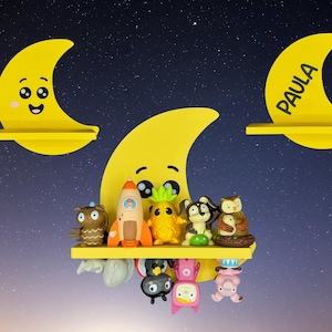 May include: Yellow crescent moon-shaped shelves with smiling faces and the name "Paula" on one. The shelves hold various colorful figurines, including an owl, rocket, pineapple, and other whimsical characters. The background is a starry night sky.