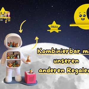 May include: A whimsical display featuring a white astronaut-shaped shelf filled with figurines, a star-shaped shelf, and a crescent moon shelf. The shelves are set against a starry night sky backdrop, with the word "Paula" on a star-shaped shelf.