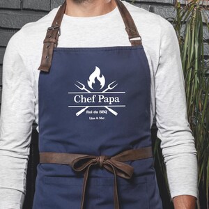 Puede incluir: Delantal azul marino con correas de cuero marrón y cinturón a juego. El delantal presenta el texto "Chef Papa" en blanco, junto con una llama y utensilios de barbacoa cruzados. Un accesorio práctico y elegante.