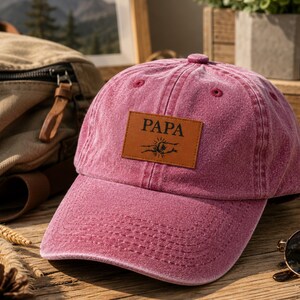 May include: A faded rose baseball cap with a leather patch displaying the word "PAPA" and a sun and airplane design. The cap rests on a wooden surface, alongside a black water bottle and sunglasses.