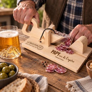 Peut inclure: Une guillotine à saucisson en bois avec une lame en métal et un cordon noir, portant les inscriptions gravées "LA SÉLECTION" et "PAPY roi de l'apéro Clément & Enzo" sur le socle. L'ustensile permet de trancher finement le saucisson sec.