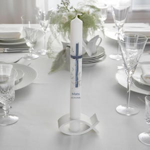 Könnte beinhalten: Eine weiße Kerze mit blauem Kreuz-Design, dem Namen "Mats" und dem Datum "02.04.2026". Sie steht in einem weißen Fischhalter auf einer weißen Tischdecke, umgeben von Gläsern und Blumen, für eine festliche Tischdekoration.