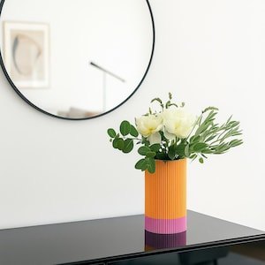 Op de afbeelding: Een moderne, kleurrijke vaas met een levendig oranje en roze gestreept ontwerp, met witte bloemen en groen blad. De vaas staat op een zwart oppervlak, met een ronde zwarte spiegel op de achtergrond.