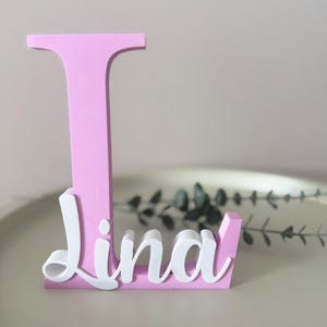 May include: A pink wooden letter "L" with the name "Lina" in white script. The letter is standing upright on a light-colored surface, with greenery in the background. The name "Lina" is attached to the base of the letter.