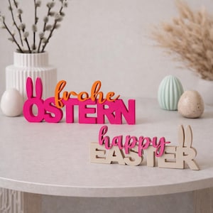 May include: Easter-themed wooden decorations. One sign reads "Frohe Ostern" in pink and orange, featuring bunny ears. Another says "happy EASTER" in pink and beige. Decorative eggs and a vase with branches complete the scene.