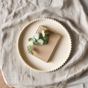 Könnte beinhalten: Ein cremefarbenes, rundes Tablett mit strukturiertem Rand hält ein braunes Buch und einen Zweig Eukalyptus. Das Tablett liegt auf einer beigen Leinenoberfläche und erzeugt eine minimalistische und natürliche Ästhetik.