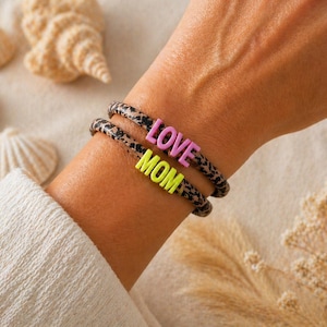 Könnte beinhalten: Zwei Armbänder mit Leopardenmuster. Eines trägt das Wort "LOVE" in rosa Buchstaben, das andere das Wort "MOM" in neongelben Buchstaben. Die Armbänder werden am Handgelenk getragen.