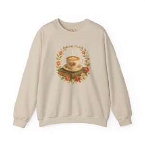 Könnte beinhalten: Cremefarbenes Sweatshirt mit floralem Design. Das Design zeigt eine Kaffeetasse mit herzförmigem Schaum, die auf einem Stapel Bücher ruht. Die Tasse ist von einem Kranz aus Rosen und Grün umgeben.