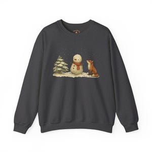 Könnte beinhalten: Dunkelgraues Sweatshirt mit einer Illustration einer Winterszene. Die Grafik zeigt einen Schneemann mit rotem Schal, einen kleinen Tannenbaum und einen Fuchs. Im Hintergrund fallen Schneeflocken. Das Sweatshirt hat lange Ärmel und einen Rundhalsausschnitt.