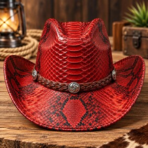 Pode incluir: Um chapéu de cowboy vermelho com um padrão texturizado de pele de cobra e detalhes em preto. O chapéu é adornado com uma faixa de couro trançado marrom decorada com três conchos metálicos prateados com um design em espiral.