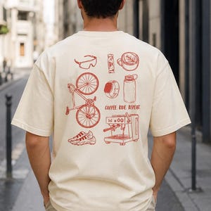 Könnte beinhalten: Ein cremefarbenes T-Shirt mit einem roten Grafikdruck auf der Rückseite. Das Design zeigt Illustrationen von einem Fahrrad, Kaffeetasse, Espressomaschine, Trinkflasche, Uhr, Sonnenbrille und Turnschuhen sowie den Text COFFEE. RIDE. REPEAT.