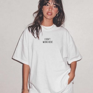 Könnte beinhalten: Ein weißes Kurzarm-T-Shirt mit dem schwarzen Text "I DON'T WORK HERE", der in einer einfachen serifenlosen Schrift mittig auf der Brust gedruckt ist. Das Kleidungsstück hat einen Rundhalsausschnitt und eine lockere Passform mit weiten Ärmeln.
