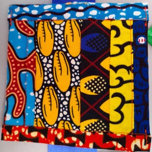 May include: A patchwork textile with a variety of bold patterns and colours. The design incorporates blue, red, yellow, and black fabrics, with geometric and organic shapes. The edges are neatly stitched with red thread, creating a textured and visually appealing piece.