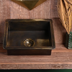 May include: A rectangular, dark bronze-colored sink with a drain. The sink is set on a wooden shelf, with a decorative vase holding dried palm leaves to the right. A geometric gold-colored accent is above the sink.