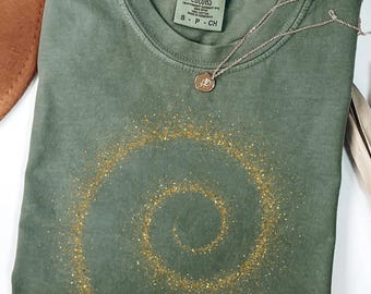 May include: A moss green Comfort Colors t-shirt with a gold glitter spiral design. A silver necklace with a pendant rests on the shirt. The shirt is folded and ready to wear.
