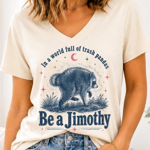 May include: A cream-colored V-neck t-shirt featuring a dark blue graphic of a raccoon in grass with the text "In a world full of trash pandas Be a Jimothy" and small pink stars and a crescent moon surrounding the central animal illustration.
