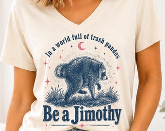 May include: A cream-colored V-neck t-shirt featuring a dark blue graphic of a raccoon in grass with the text "In a world full of trash pandas Be a Jimothy" and small pink stars and a crescent moon surrounding the central animal illustration.