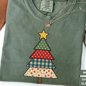 May include: A moss green short-sleeve t-shirt featuring a patchwork Christmas tree design. The tree has a yellow star on top and is made of various patterned fabrics, including polka dots, gingham, and floral prints. A silver necklace with a pendant is also visible.