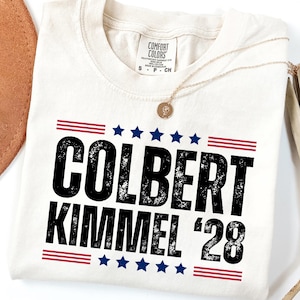 May include: Cream-colored t-shirt with the text "COLBERT KIMMEL '28" in black, framed by red and blue stripes and stars. A gold necklace with a pendant rests on the shirt. The shirt is folded on a white surface.