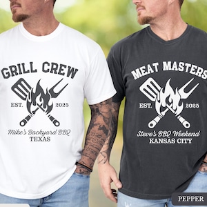 May include: Two t-shirts, one white and one pepper-colored, featuring barbecue-themed designs. The white shirt reads "GRILL CREW" with "Mike's Backyard BBQ TEXAS" below. The pepper shirt says "MEAT MASTERS" with "Steve's BBQ Weekend KANSAS CITY". Both shirts have a grill graphic.