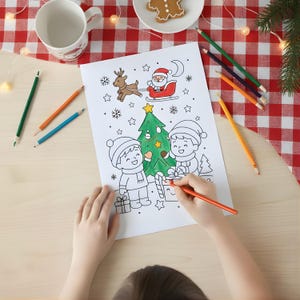 Child coloring a Christmas tree scene with Santa and reindeer using colored pencils on a festive table with a red checkered tablecloth.