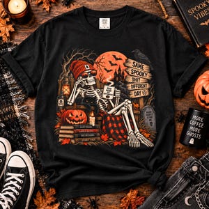 May include: A black t-shirt featuring a graphic of a skeleton wearing a red beanie and sunglasses, sitting in a chair with a mug that says Dead Inside But Caffeinated, a pumpkin, books, and a sign that reads Same Spooky Shit Different Day.