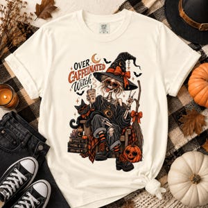 May include: An off-white t-shirt featuring a graphic of a skeleton wearing a witch hat and heart-shaped sunglasses, holding a coffee cup. The text reads Over Caffeinated Witch. A black cat, books, a broom, and a pumpkin are also depicted in the design.