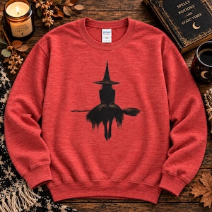 May include: A heathered red crewneck sweatshirt featuring a black graphic print of a silhouette of a witch wearing a pointed hat and sitting on a broomstick. The garment has long sleeves, a ribbed collar, and a Gildan brand label at the inner neckline.