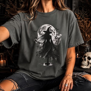 May include: A charcoal gray short-sleeve t-shirt featuring a graphic print of a witch in a pointed hat standing before a large full moon. The design includes silhouettes of flying bats, swirling clouds, and a reflection on the ground below.