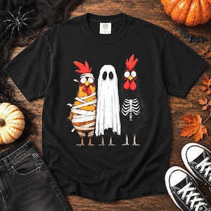 Op de afbeelding: Een zwart t-shirt met korte mouwen en een afbeelding van drie kippen in Halloween-kostuums. Eén kip is ingepakt als een mummie, één draagt een wit laken als spook en de derde is verkleed als skelet. Het shirt heeft een ronde hals.