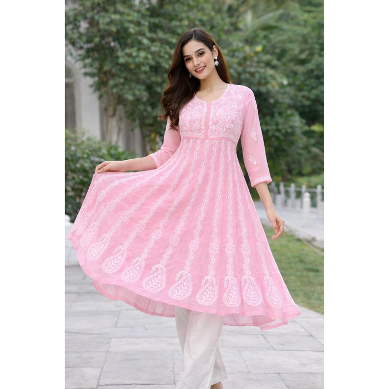 May include: A light pink, embroidered tunic dress with a flared skirt and three-quarter sleeves. The dress features intricate white floral embroidery and is paired with white pants. The woman is smiling and standing outside.
