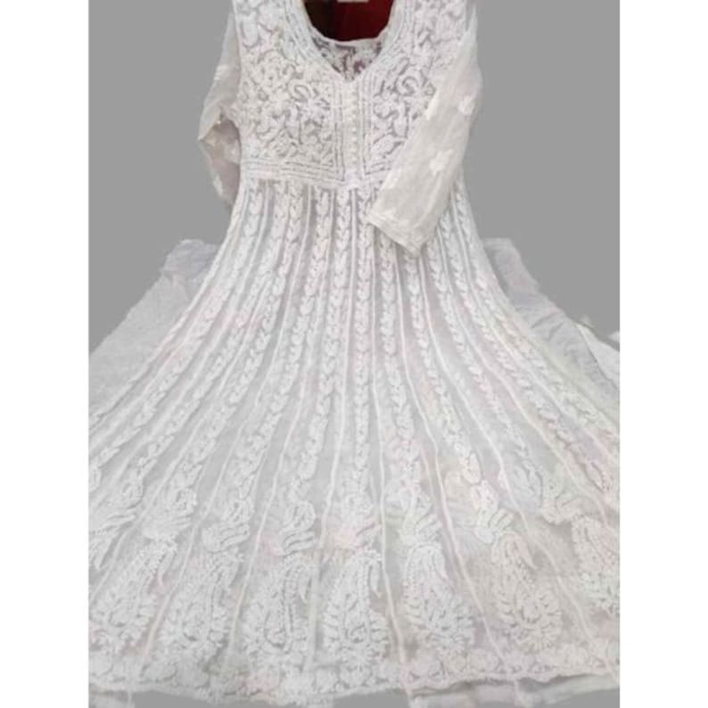 May include: A white, long-sleeved dress with intricate embroidered detailing. The dress features a V-neckline and a flowing skirt, showcasing a delicate, lacy pattern. The embroidery is visible across the bodice and sleeves, creating a textured, elegant look.