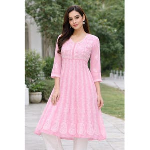May include: A light pink, embroidered tunic dress with a V-neck and three-quarter sleeves. The dress features intricate white embroidery and a flared skirt, paired with white pants. The outfit is suitable for casual or semi-formal occasions.