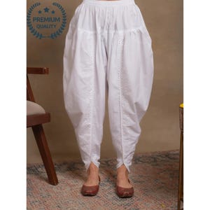 Puede incluir: Pantalones salwar de algodón blanco con cintura fruncida y tobillos cónicos. Los pantalones presentan costuras decorativas en la parte delantera y en los laterales. La imagen incluye un sello de "Premium Quality".