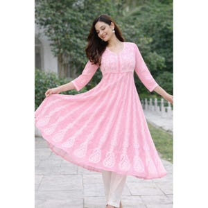 May include: A light pink, embroidered tunic dress with a flared skirt and three-quarter sleeves. The dress features white embroidery and is paired with white pants. The dress has a V-neckline and a flowing silhouette.