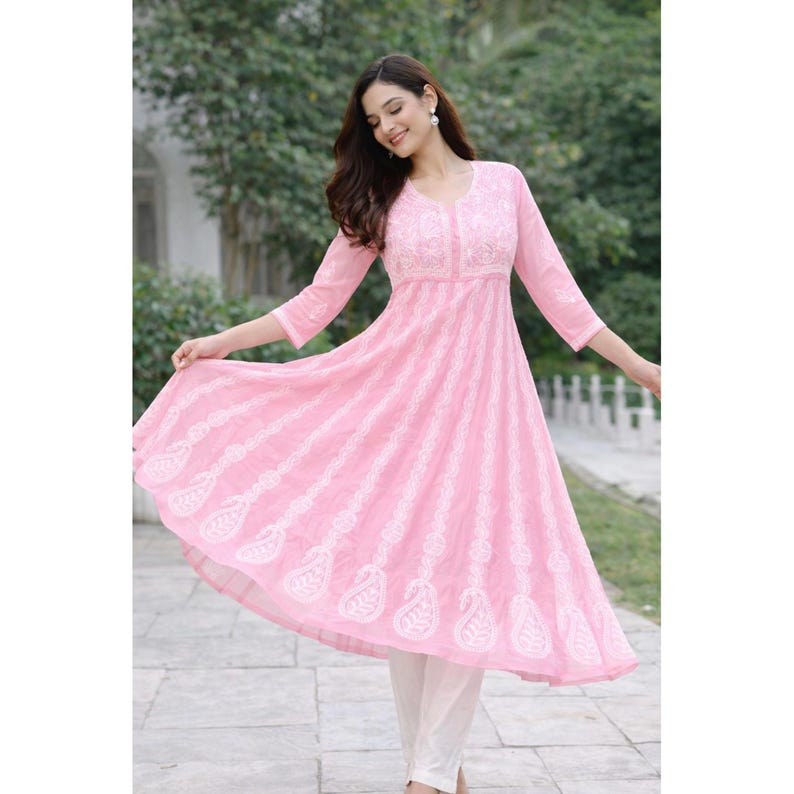 May include: A light pink, embroidered tunic dress with a flared skirt and three-quarter sleeves. The dress features white embroidery and is paired with white pants. The dress has a V-neckline and a flowing silhouette.