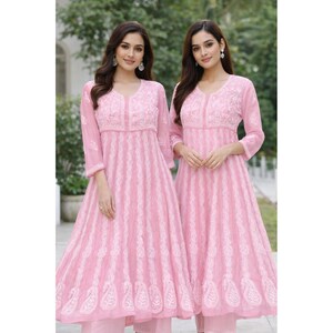 May include: Two women wearing matching pink embroidered dresses. The dresses have a V-neck, three-quarter sleeves, and a flared skirt. The dresses are adorned with white embroidery and are paired with matching pink pants. The background is a blurred outdoor setting.