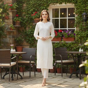 Può includere: Una donna indossa un kurta bianco ricamato e pantaloni abbinati. Il kurta ha uno scollo tondo e maniche a tre quarti. L'outfit è indossato all'aperto, davanti a un muro di pietra ricoperto di verde e fiori.