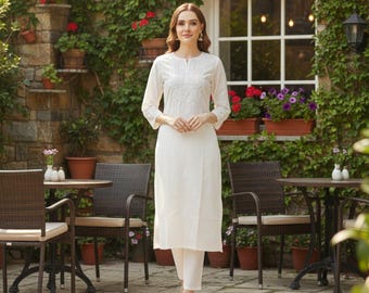 May include: A woman in a white embroidered kurta and trousers. The kurta features a round neck and three-quarter sleeves. The outfit is worn outside, against a stone wall covered in plants and flowers, near a window.