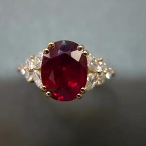 May include: A gold ring with a large, oval-cut ruby at its centre. The ruby is a deep red hue, encircled by smaller, clear gemstones. The ring showcases a classic, elegant design.