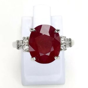 May include: A silver ring featuring a large, oval-cut ruby gemstone. The ruby is a deep red color and is flanked by smaller, clear gemstones on each side. The ring is displayed on a white stand.