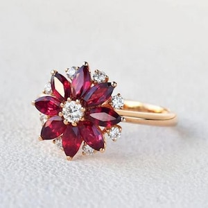 Peut inclure: Bague en or avec un motif floral. Elle présente des pétales rouge foncé et de petites pierres blanches, avec une pierre blanche plus grande au centre. L'anneau est posé sur un fond doux et neutre.